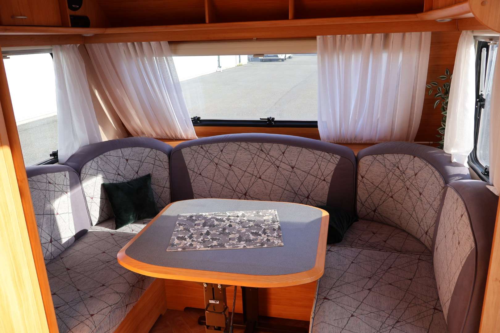 Ett sittutrymme i en husvagn med U-formad soffa klädd i grått tyg med mönster, matchande ryggstöd och ett rektangulärt bord i mitten med träkant och blågrå yta. Ljusa gardiner hänger framför fönstren och överskåp i trä syns ovanför.