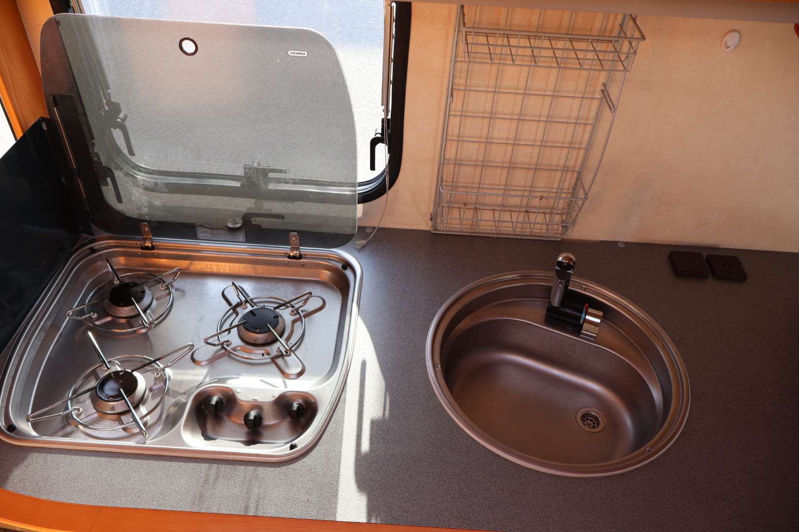 Ett kompakt kök i en husvagn med trelågig gasolspis till vänster och en rund rostfri diskho med blandare till höger. Ovanför bänken finns ett trådställ för förvaring och ett fönster med öppningsbar ruta.