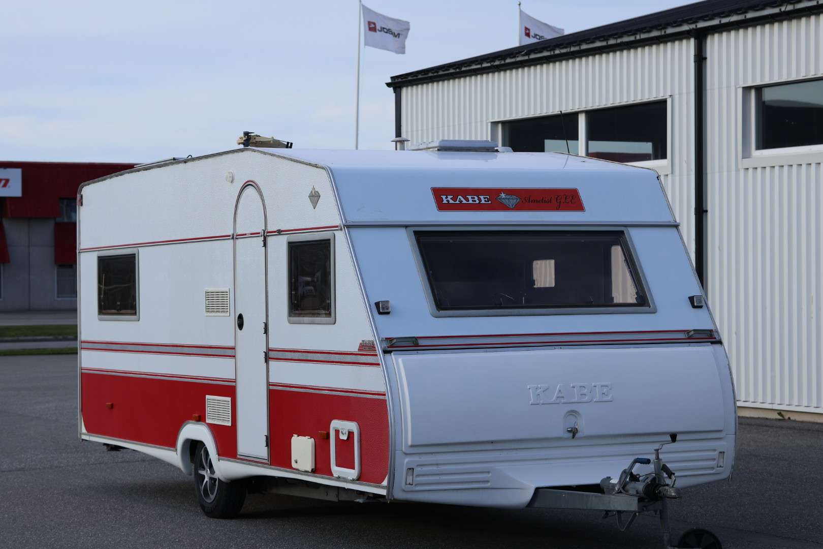 En rödvit KABE husvagn av modellen "Ametist" står parkerad utanför en industribyggnad med vita fasader.